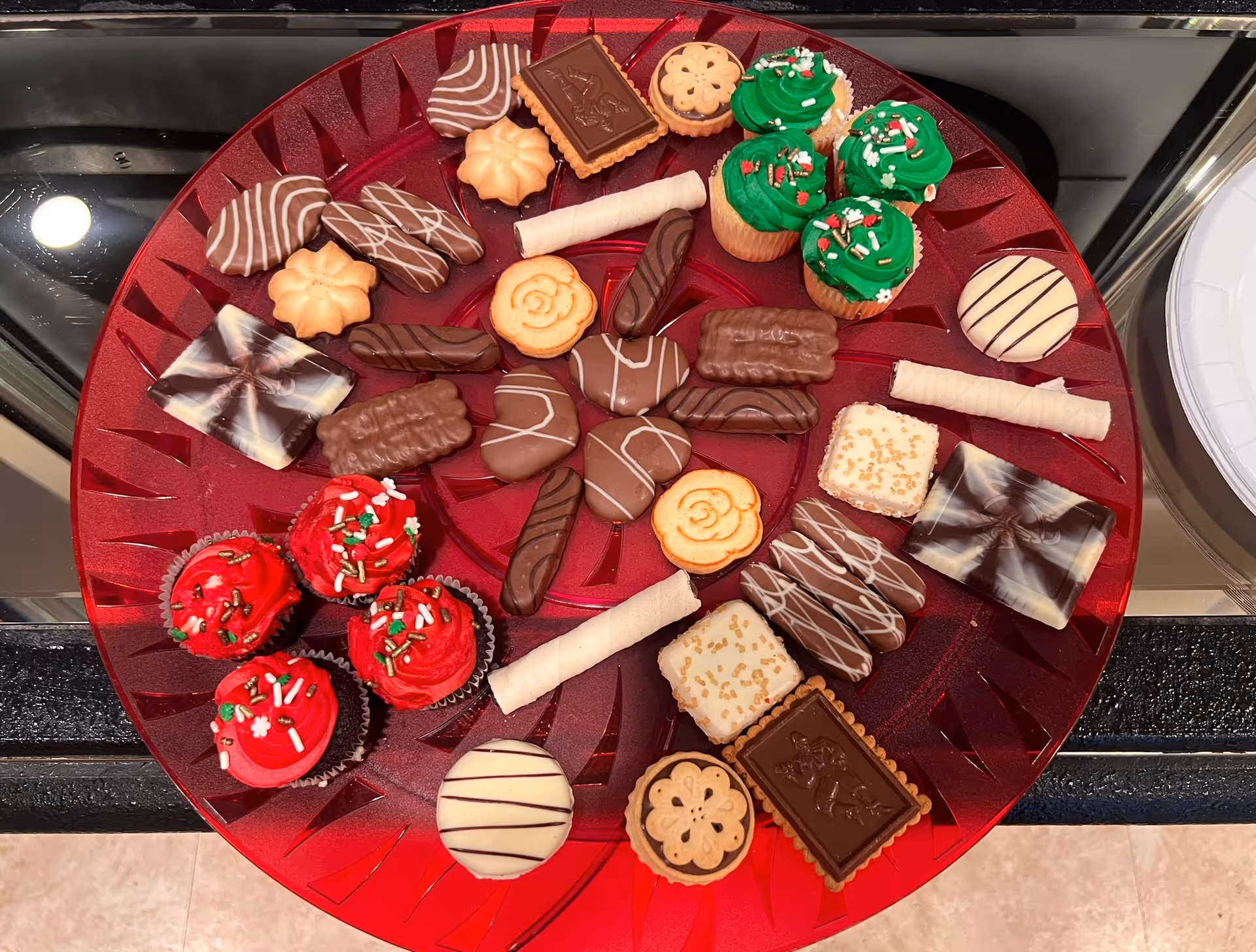 A red platter filled with an assortment of decorated cookies and mini cupcakes with red and green frosting, placed on a kitchen counter.