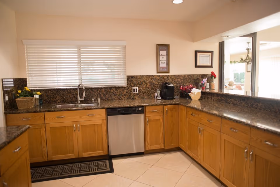 A kitchen with wooden cabinets, granite countertops, a stainless steel dishwasher, and a sink under a window with white blinds. There are decorative items including a basket with yellow flowers, a coffee maker, and framed pictures on the wall. A pass-through window opens to an adjacent room.