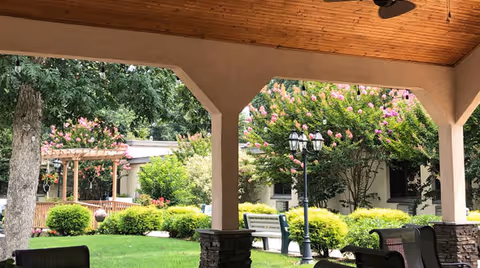 View from a covered patio area looking out onto a well-maintained garden with green grass, bushes, flowering trees, a wooden pergola, benches, and a lamp post. The patio has a wooden ceiling and stone pillars.