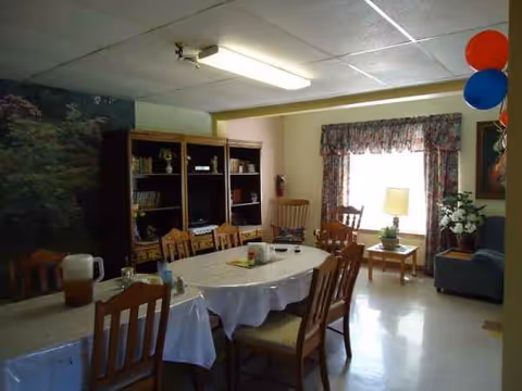 A nursing home dining room with round table, chairs, bookshelf, and balloons.