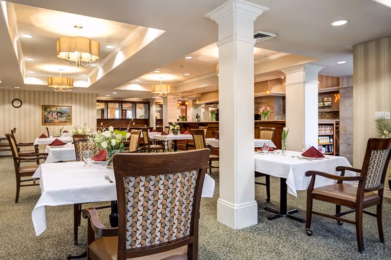 A well-lit dining room in a senior living facility with tables covered in white tablecloths, each set with folded burgundy napkins, glassware, and floral centerpieces. The room features patterned carpet, wooden chairs with cushioned seats and backs, decorative ceiling lights, and a service counter in the background.