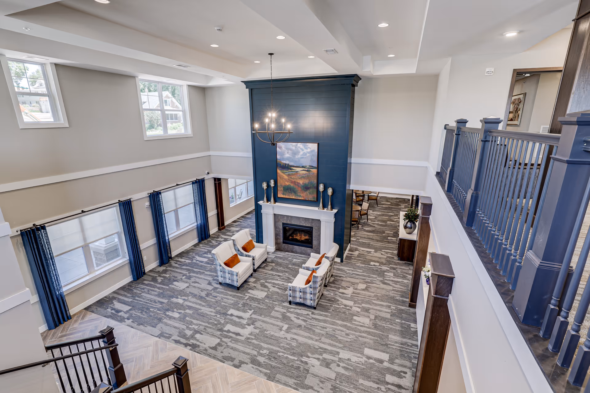 Spacious and modern living room area in a senior living facility with high ceilings, large windows with blue curtains, a central fireplace with a dark blue accent wall, four armchairs with orange cushions arranged around the fireplace, and a chandelier hanging above.