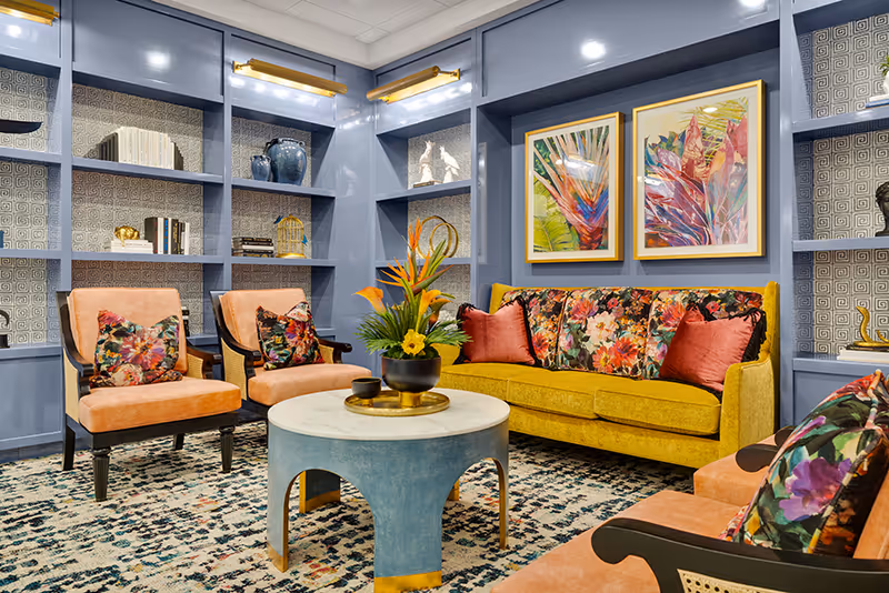 Colorful lounge with a yellow sofa, coral armchairs, blue built-in shelves, a round coffee table and floral artwork and pillows.