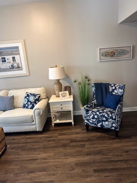 Cozy living room with a white sofa, blue patterned armchair, small white side table with a lamp, and wall art on a light gray wall.