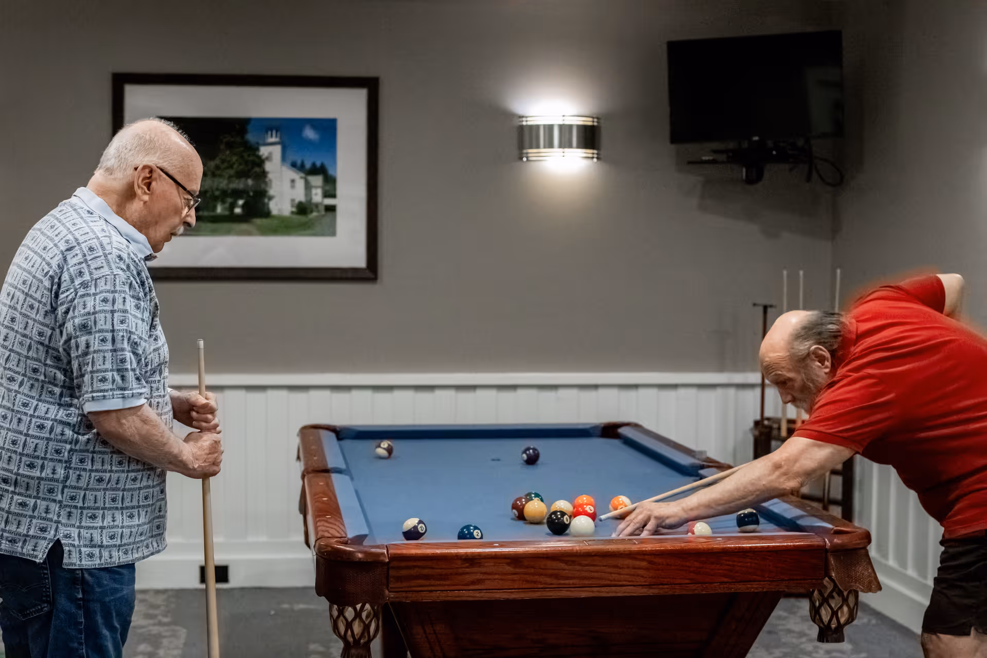 Two elderly men playing pool in a recreational room with a blue pool table. One man in a patterned shirt is standing and holding a cue stick, while the other man in a red shirt is leaning over the table aiming a shot. The room has gray walls, a framed picture, a wall light, and a mounted television.