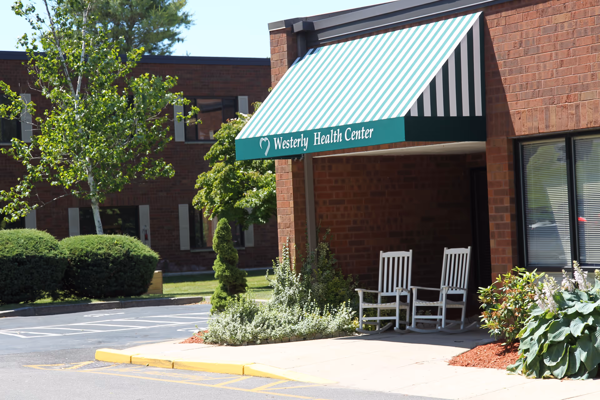 Entrance to Westerly Health Center with a green and white striped awning, two white rocking chairs on the porch, surrounded by greenery and bushes, with a brick building in the background.