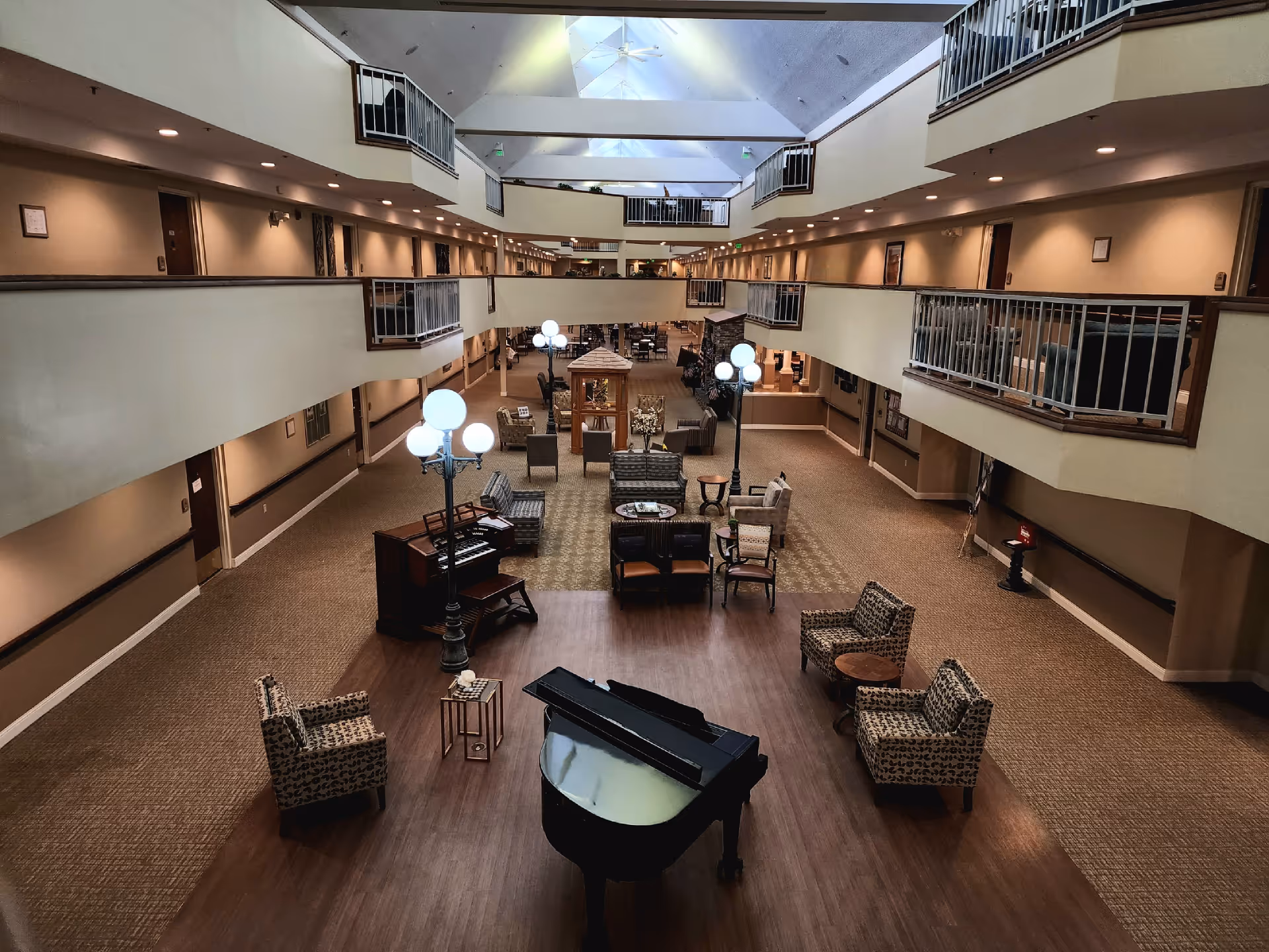 Spacious interior common area of a senior living facility with multiple seating arrangements including armchairs and sofas, a grand piano, a smaller piano, and several standing lamps with globe lights. The area has a high ceiling with skylights and a second-floor balcony overlooking the space.