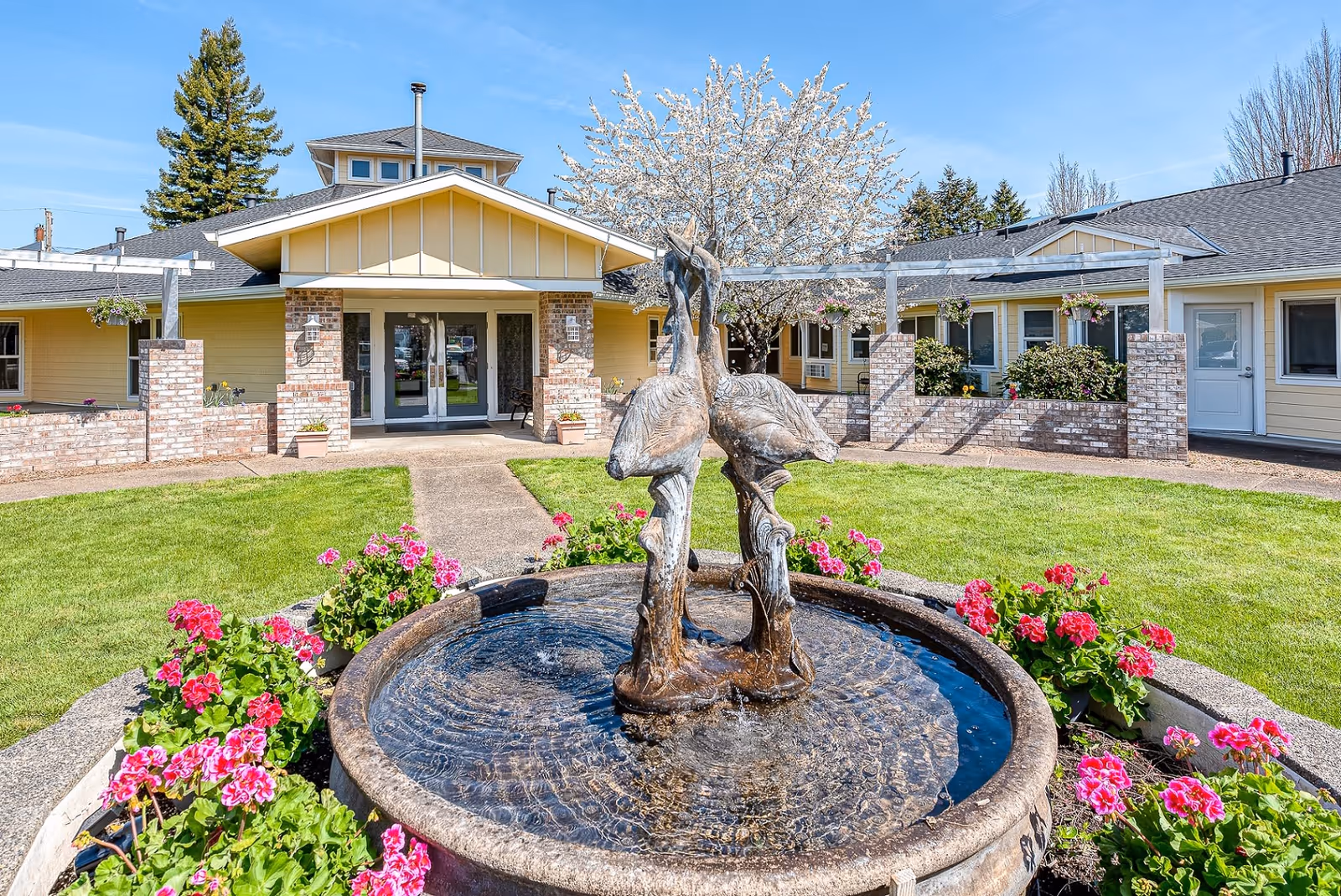 Outdoor courtyard area of Forest Grove Beehive Assisted Living featuring a round water fountain with two bird sculptures in the center, surrounded by pink flowers and green grass. The building with yellow siding and brick accents is visible in the background under a clear blue sky.