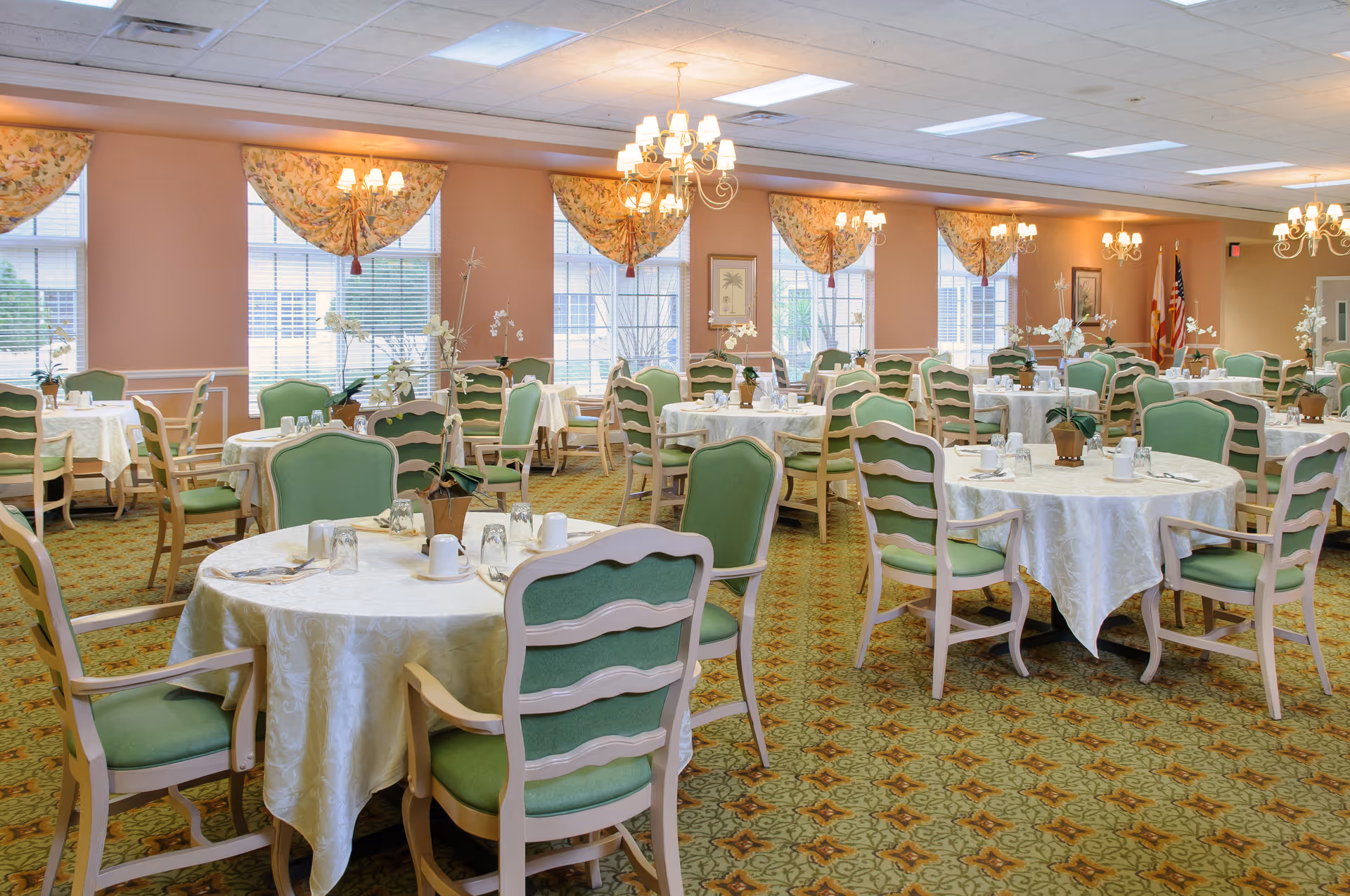 A spacious dining room with round tables covered in white tablecloths, each set with glasses, cups, and silverware. The chairs have green cushions and light wooden frames. The room has large windows with floral valances, chandeliers hanging from the ceiling, and a patterned carpet in green and brown tones.