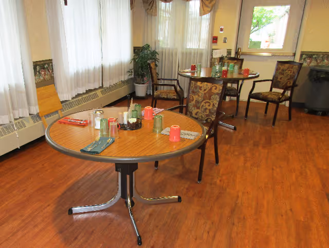A dining area with two round wooden tables set with colorful plastic cups, napkins, and condiments. The room has wood flooring, large windows with sheer white curtains, and several patterned chairs around the tables. A potted plant is placed near the windows.