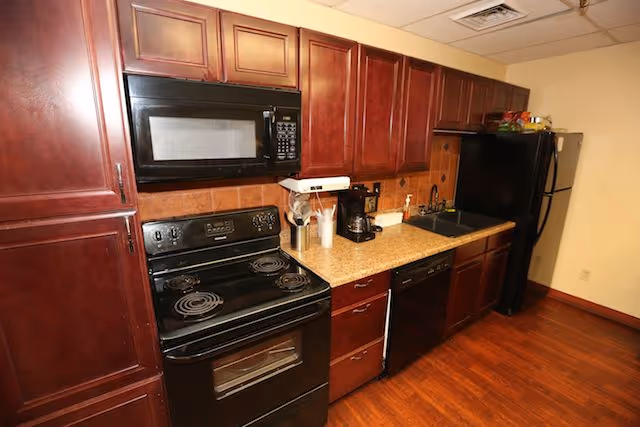 Small kitchen with dark wood cabinets, black stove and microwave, refrigerator and dishwasher, and a countertop with sink and coffee maker.