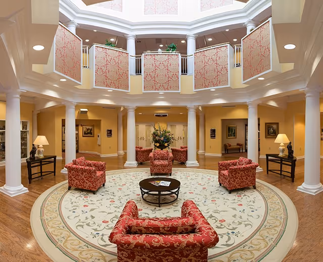 A spacious, elegant circular lounge area with a high ceiling and a skylight. The room features white columns surrounding the space, patterned red armchairs arranged around a round coffee table on a large decorative rug. The walls are painted a warm yellow, and there are side tables with lamps and decorative statues. Upstairs balcony railings have red patterned fabric panels, and there are framed artworks on the walls.