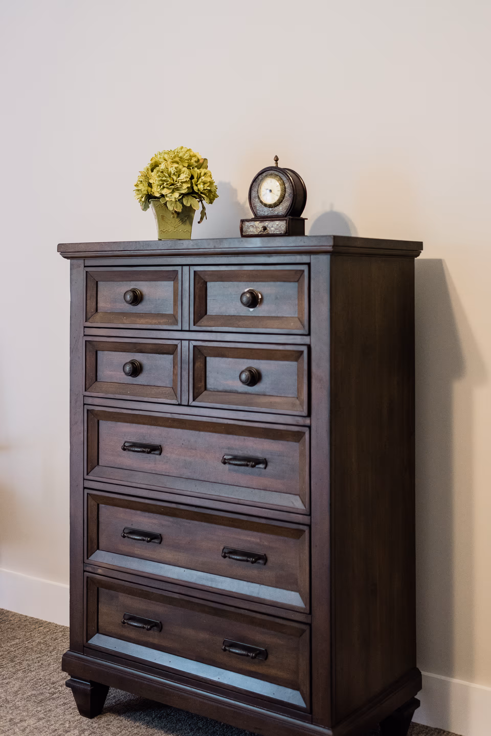 A dark wooden chest of drawers with six drawers, placed against a beige wall on a carpeted floor. On top of the chest, there is a small green potted plant with flowers and a vintage-style round clock.