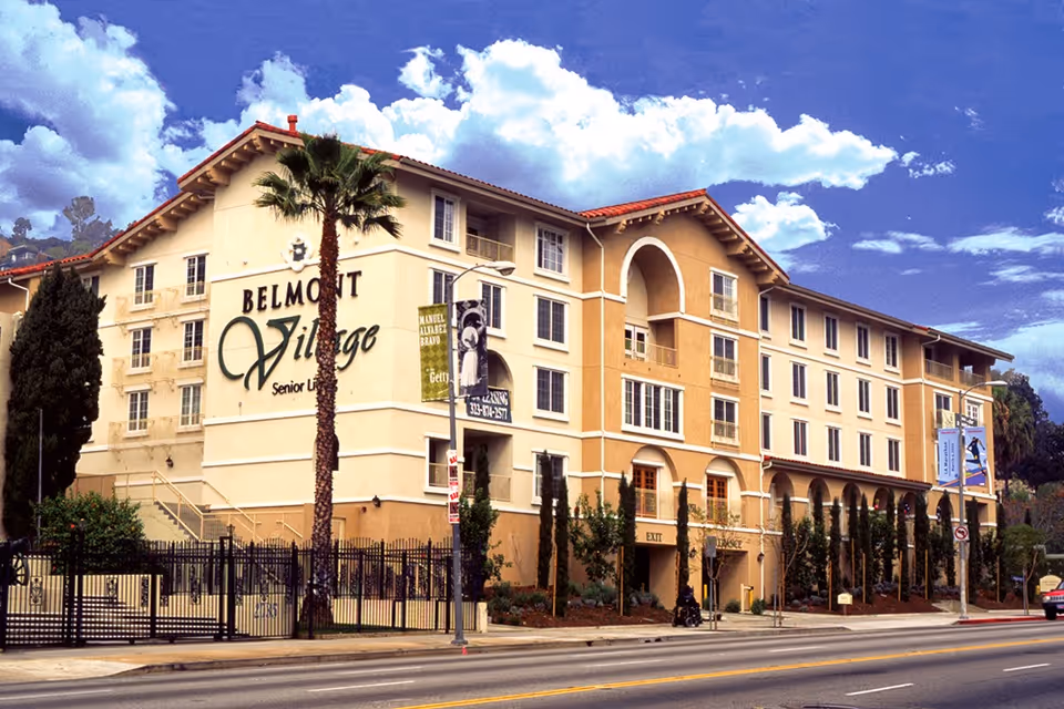 Exterior view of Belmont Village Senior Living Hollywood Hills, a multi-story building with beige and light brown walls, several windows, and a red-tiled roof. There are palm trees and other greenery in front of the building, a black fence along the sidewalk, and a clear blue sky with some clouds above.