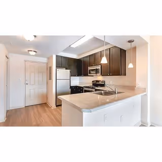 Modern kitchen area with light-colored countertops, dark wood cabinets, stainless steel refrigerator, stove, microwave, and a double sink. The kitchen features pendant lighting and a wooden floor. A door and hallway are visible in the background.