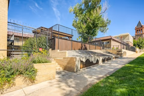 Outdoor view of a senior living facility with a paved walkway, green grass, a tree, and a fenced area with brick retaining walls under a clear blue sky.