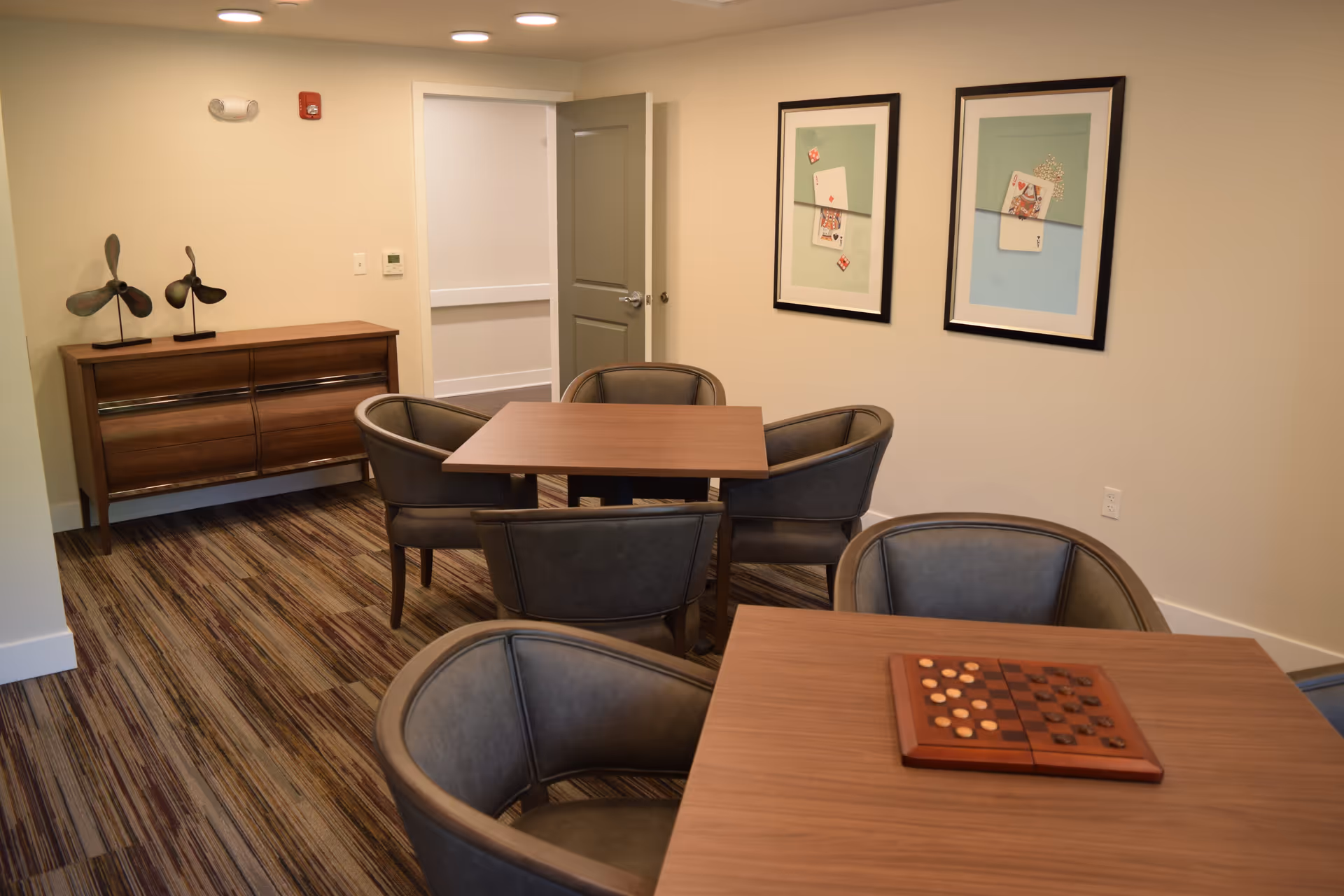 A small room with two square wooden tables, each surrounded by four cushioned chairs. One table has a wooden checkers game set on it. The room has a striped carpet, beige walls, and two framed pictures of playing cards and dice on the wall. A wooden sideboard with two decorative metal sculptures is against one wall, and an open door leads to a hallway.