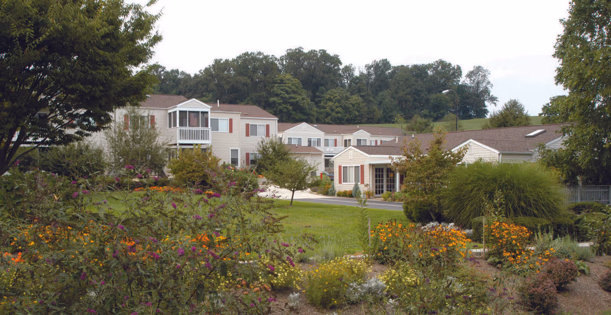 View of a retirement community with beige buildings featuring red shutters, surrounded by lush green lawns, colorful flower beds, and trees under a cloudy sky.