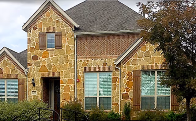 Stone-faced building front with multiple windows, wooden shutters, and landscaping.