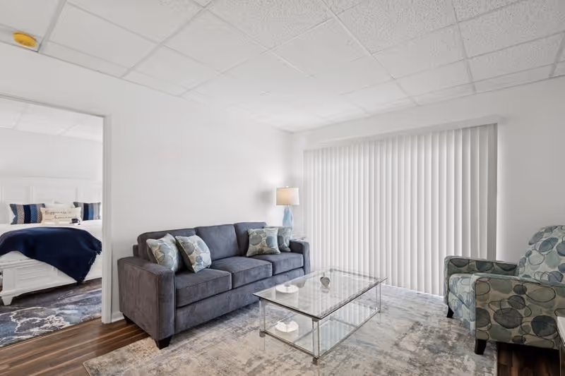 A bright living room with a gray sofa adorned with patterned cushions, a glass coffee table, a patterned armchair, a floor lamp, and vertical blinds covering a large window. A bedroom with a white bed and blue bedding is visible through an open doorway.