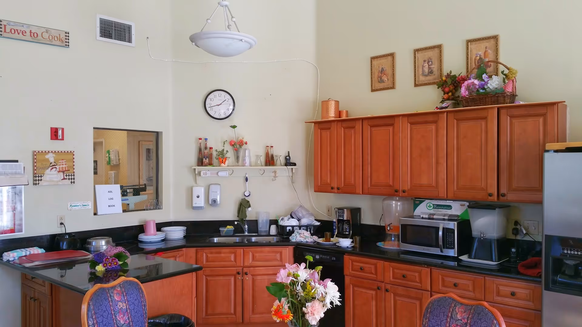 A kitchen area with wooden cabinets, a black countertop, a microwave, a refrigerator, and various kitchen items including plates, cups, and flowers. There is a clock on the wall, decorative pictures, and a sign that says 'Love to Cook'.