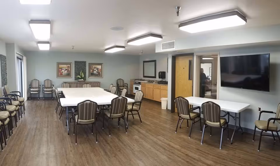 Large multipurpose dining/activity room with folding tables and chairs, a wall-mounted TV, and a small kitchenette.