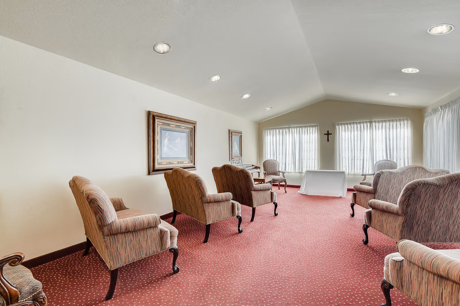 A spacious room with red carpet and several upholstered armchairs arranged in two rows facing a small table covered with a white cloth near large windows with sheer white curtains. The walls are light-colored with framed artwork and a small cross hanging between the windows. The ceiling has recessed lighting.