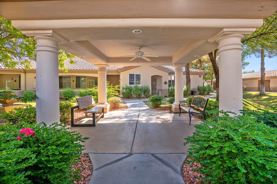 Covered outdoor seating area with two benches under a roof supported by large white columns, surrounded by green bushes and trees, with a building in the background at The Montecito Senior Living.