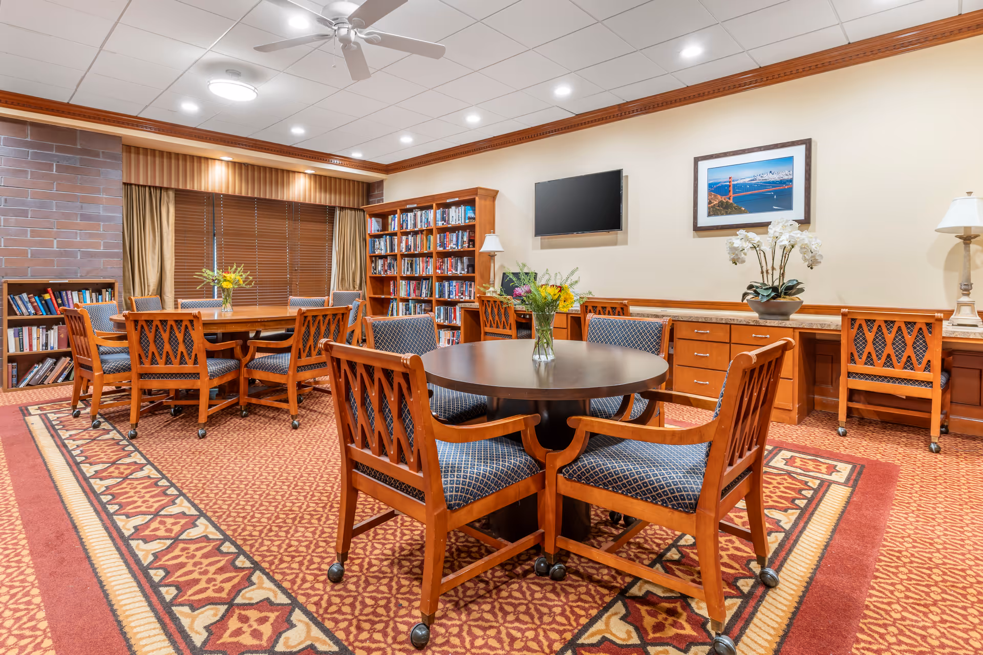 A cozy common area with wooden tables and chairs featuring blue patterned cushions. There are two flower vases on the tables, a bookshelf filled with books, a flat-screen TV mounted on the wall, and a framed picture of the Golden Gate Bridge. The room has a patterned carpet, beige walls, and a ceiling fan with recessed lighting.