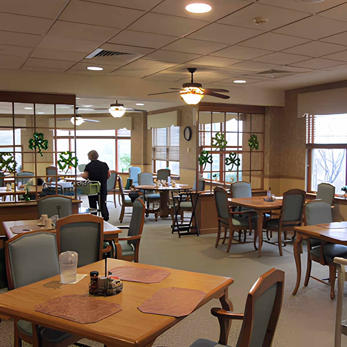 Interior view of a dining room in a senior living facility with multiple wooden tables and cushioned chairs. A person is seen walking away in the background. The room has large windows with blinds, ceiling fans with lights, and decorative green shamrocks on the glass partitions.