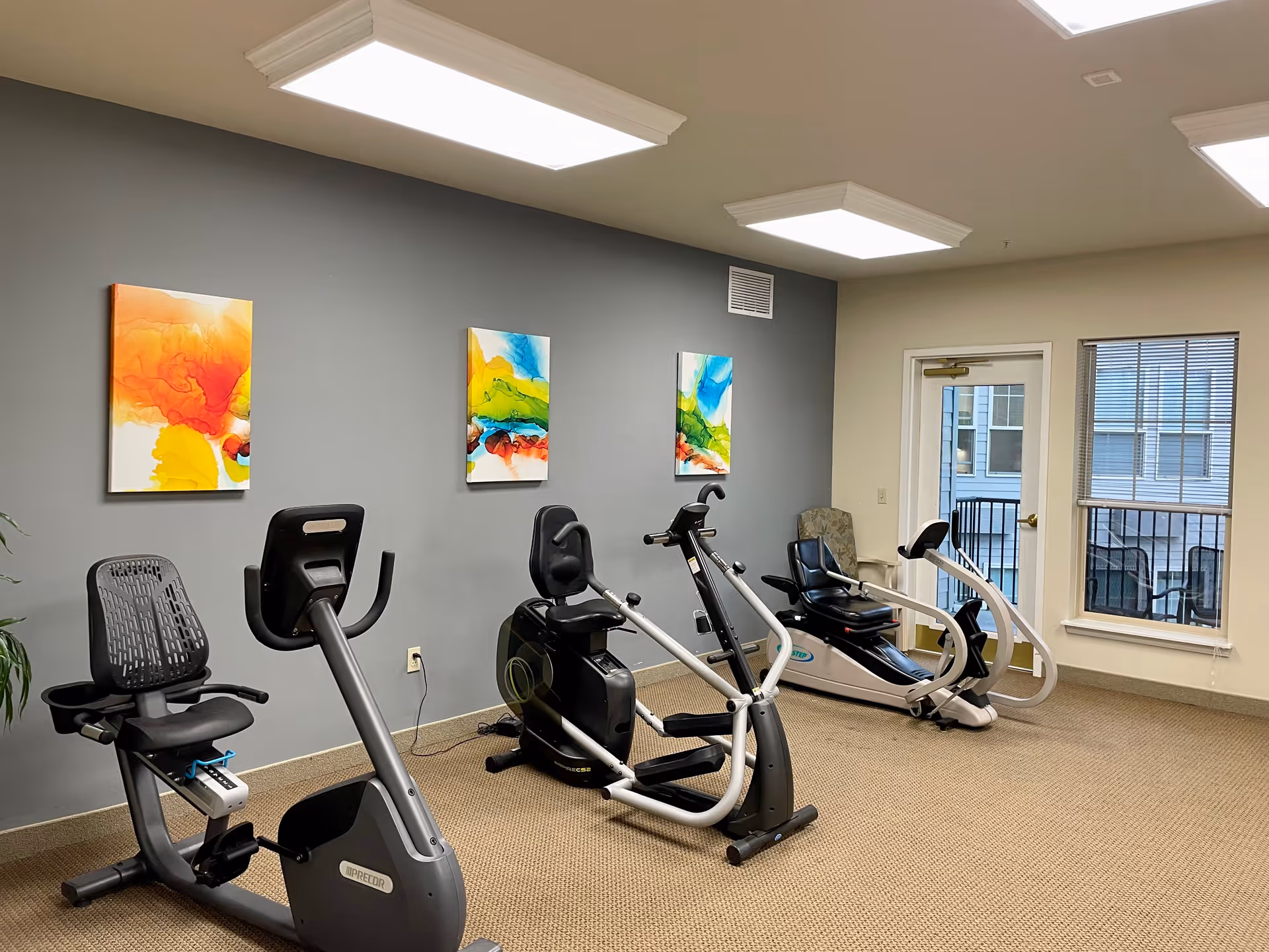 A small fitness room with three exercise machines including two recumbent bikes and one rowing machine. The room has gray walls with three colorful abstract paintings hanging on one wall. There is a window and a glass door leading to an outdoor balcony. The floor is carpeted and the ceiling has bright rectangular lights.