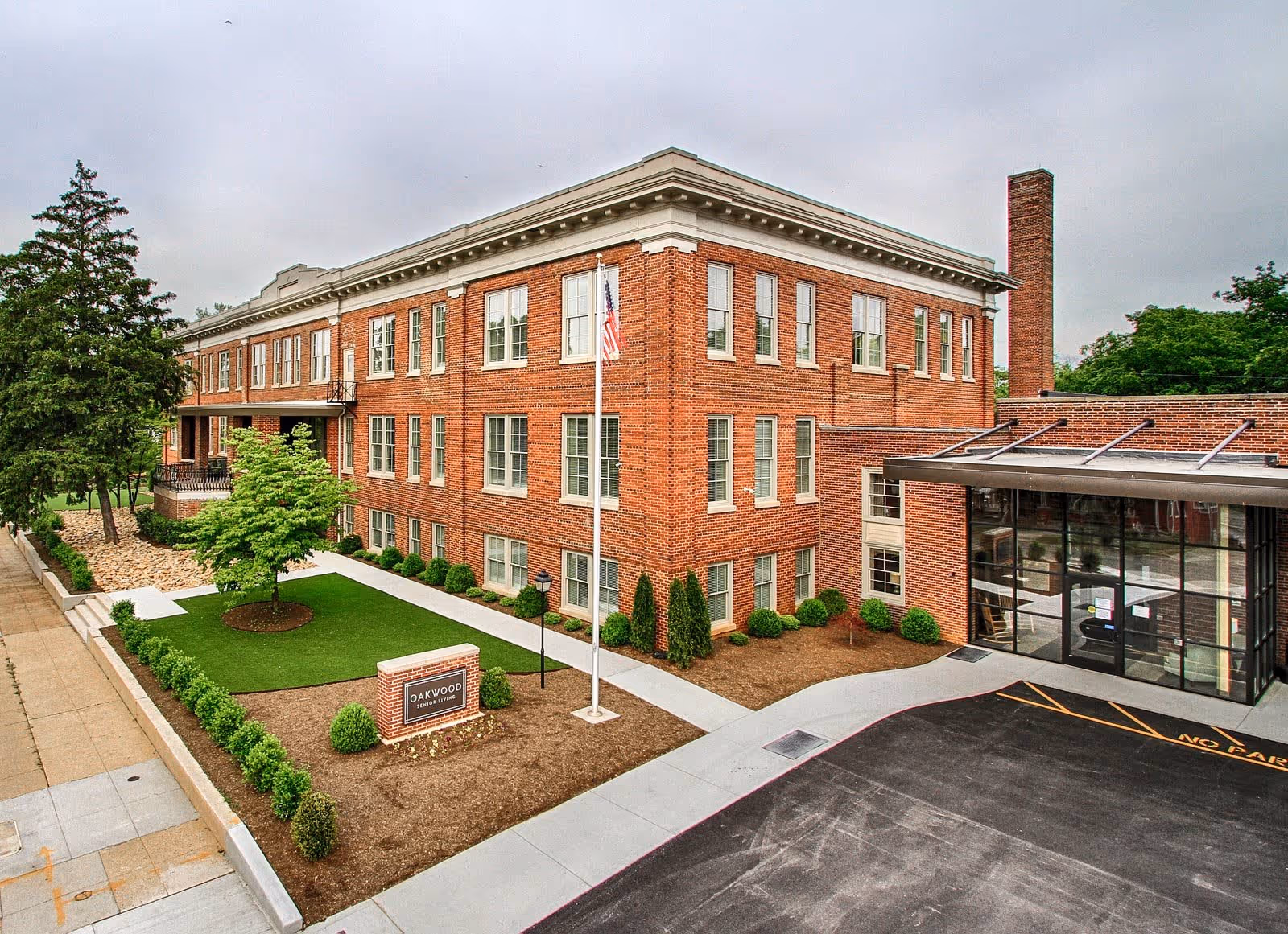 Exterior view of a three-story brick building with multiple windows, a small landscaped lawn with a tree and bushes, a flagpole with an American flag, and a driveway leading to a glass entrance. A sign in front reads 'Oakwood Senior Living'.
