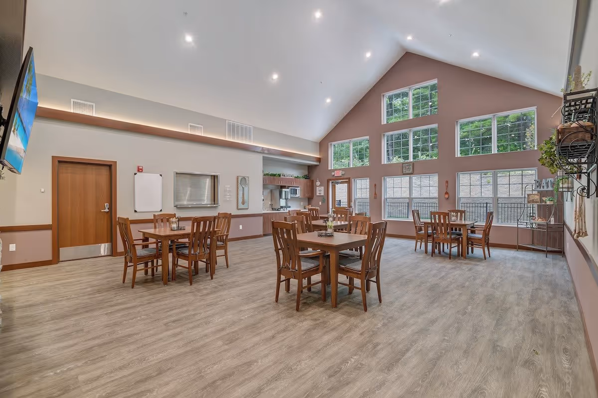 A spacious dining room with several wooden tables and chairs arranged neatly. The room features large windows with multiple panes allowing natural light to fill the space. The walls are painted in neutral tones with decorative elements such as a large spoon and fork artwork and a metal shelving unit with plants and decor. The ceiling is vaulted with recessed lighting, and a flat-screen TV is mounted on one wall. The floor has a light wood finish.