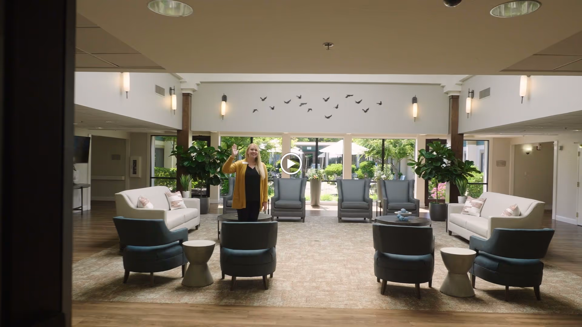 A spacious and well-lit senior living facility common area with comfortable seating including sofas and armchairs arranged around small tables. A woman in a yellow cardigan is standing and waving in the center of the room. Large windows at the back provide a view of a green outdoor garden area. Wall-mounted lights and decorative bird wall art are visible above the windows.
