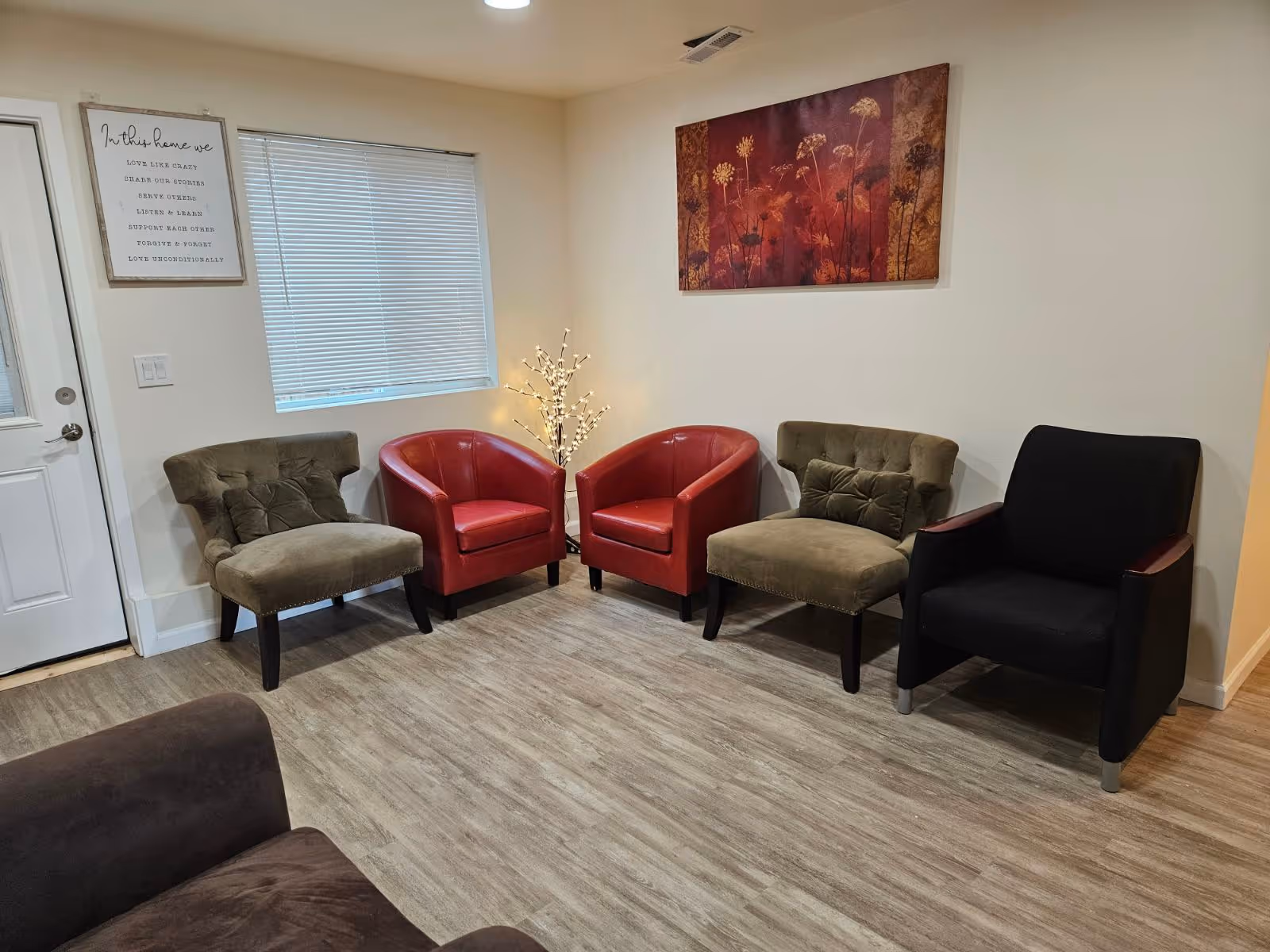 A cozy seating area with two red armchairs, two olive tufted chairs and a black armchair arranged along a wall with artwork and a small lit decorative tree.