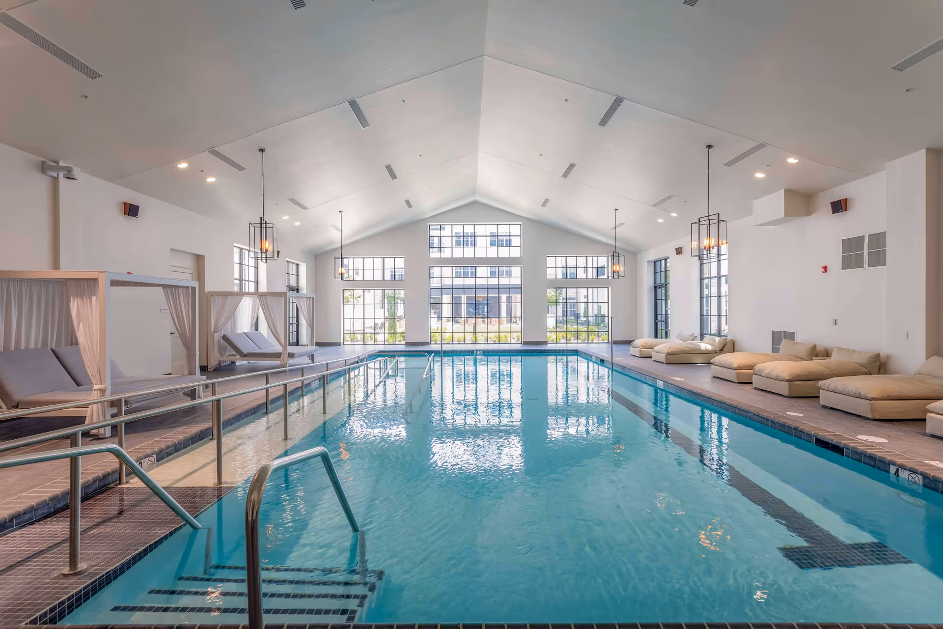 Indoor swimming pool with clear blue water, metal handrails, and steps leading into the pool. The pool area features large windows letting in natural light, cushioned lounge chairs on the right side, and canopy-covered lounge beds on the left side. The ceiling is vaulted with modern light fixtures hanging down.