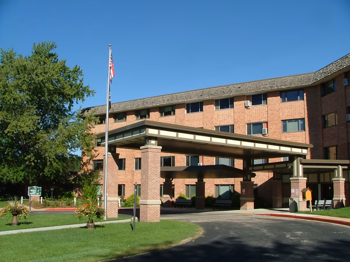 Front entrance of a brick multi-story retirement community with a covered porte-cochère and an American flag on a flagpole.