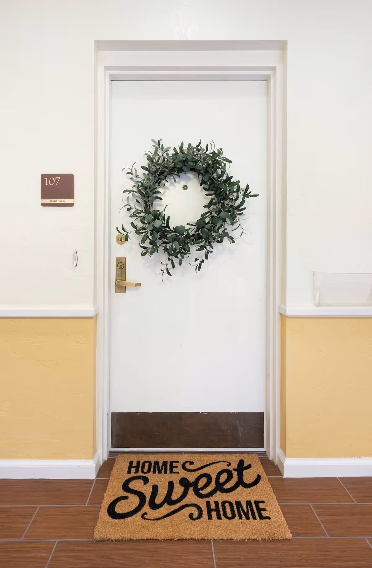 White door with a green leafy wreath hanging on it, a brass door handle and peephole. To the left of the door is a sign with the number 107 and the label 'Model Room'. A doormat in front of the door reads 'Home Sweet Home'. The walls are painted white on the upper half and yellow on the lower half, with brown tiled flooring.
