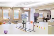 A spacious rehabilitation room with parallel bars for walking exercises, colorful therapy balls, cones for obstacle courses, and various exercise equipment. The room has large windows with blinds, light-colored walls, and a clean, bright atmosphere.