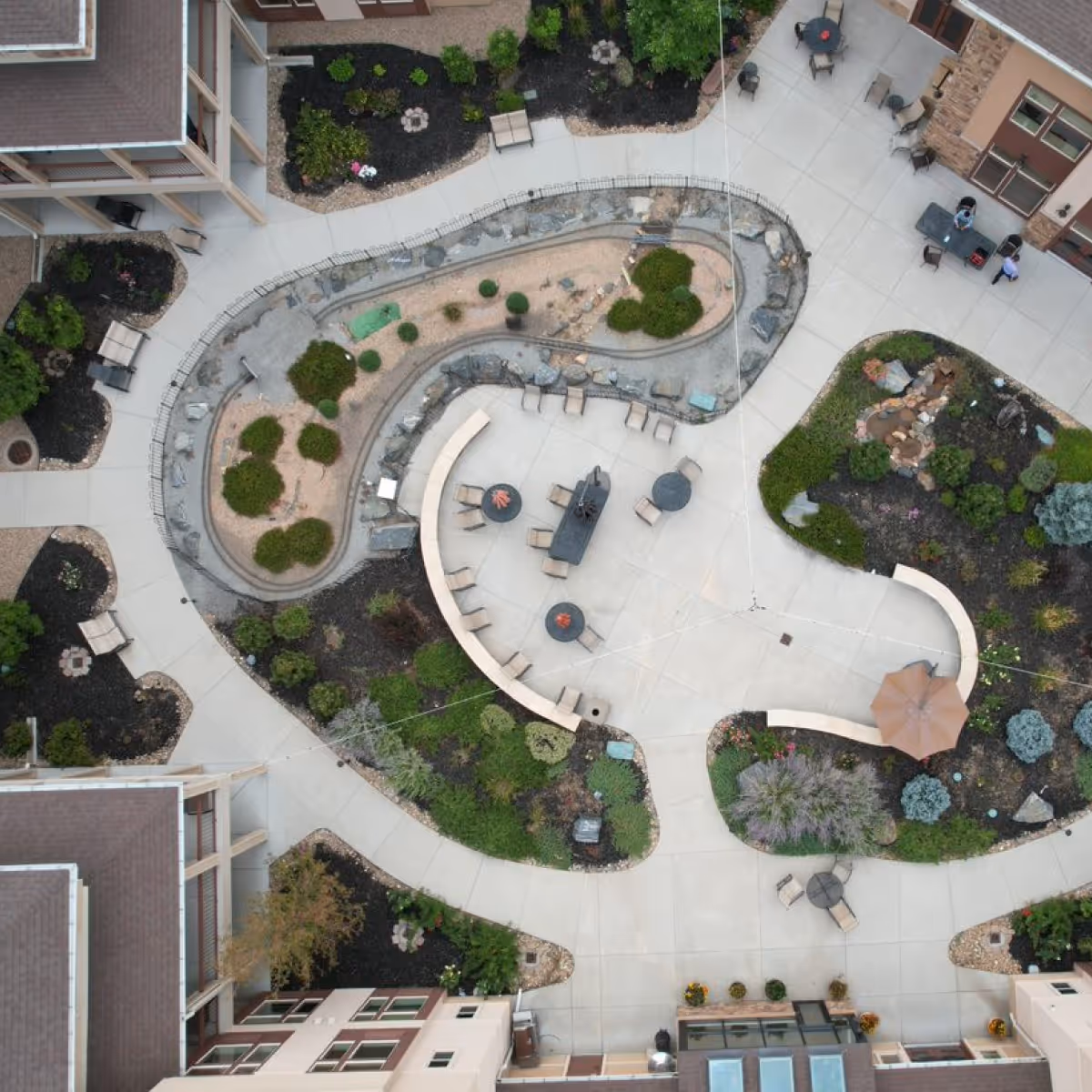 Aerial view of an outdoor courtyard area at a senior living facility with curved walkways, landscaped garden beds, seating areas with tables and chairs, and a large umbrella providing shade.