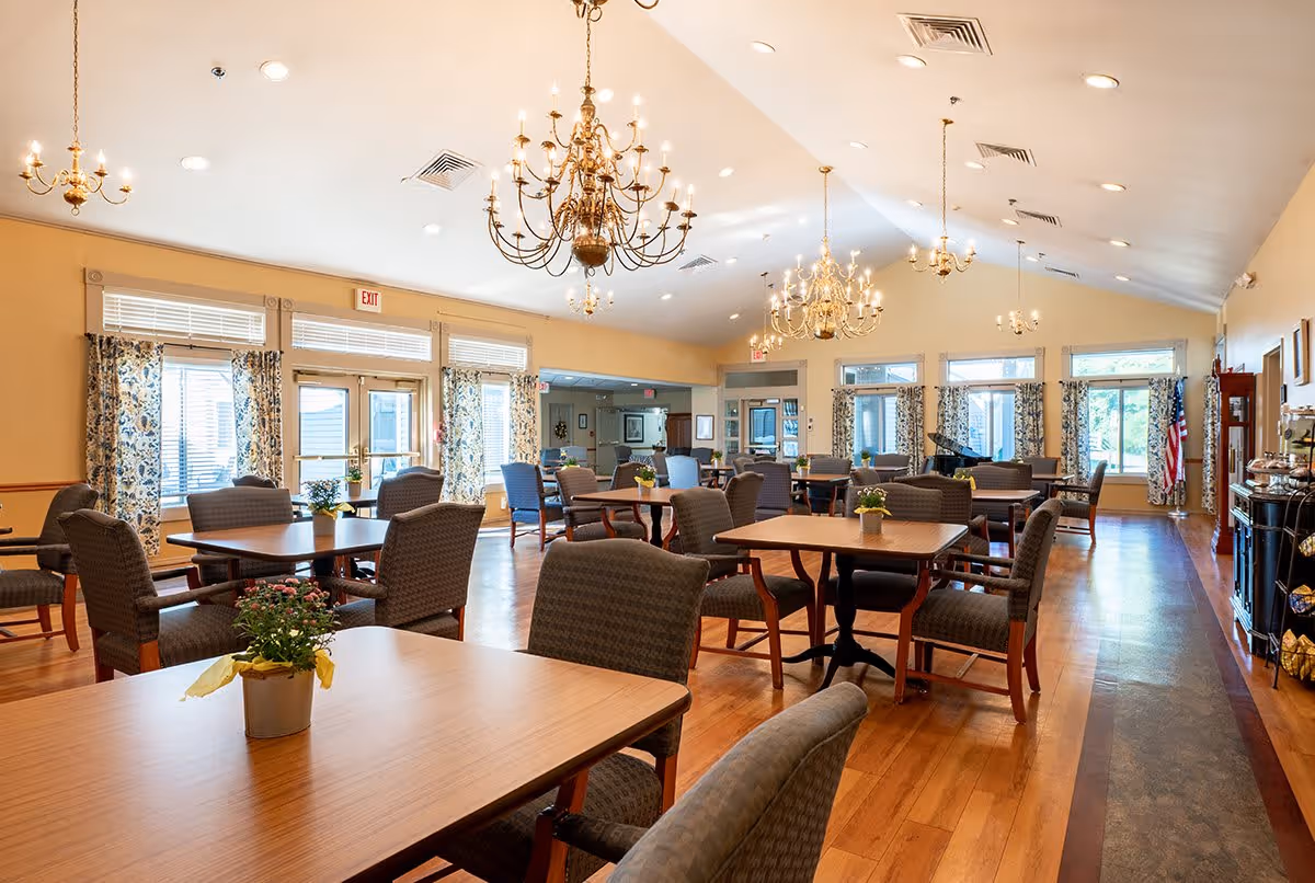 A spacious dining room with multiple wooden tables and cushioned chairs arranged neatly. The room features large windows with patterned curtains allowing natural light to fill the space. Elegant chandeliers hang from the ceiling, and there are small potted plants on the tables. The floor is wooden, and there is an American flag in the corner near a piano.