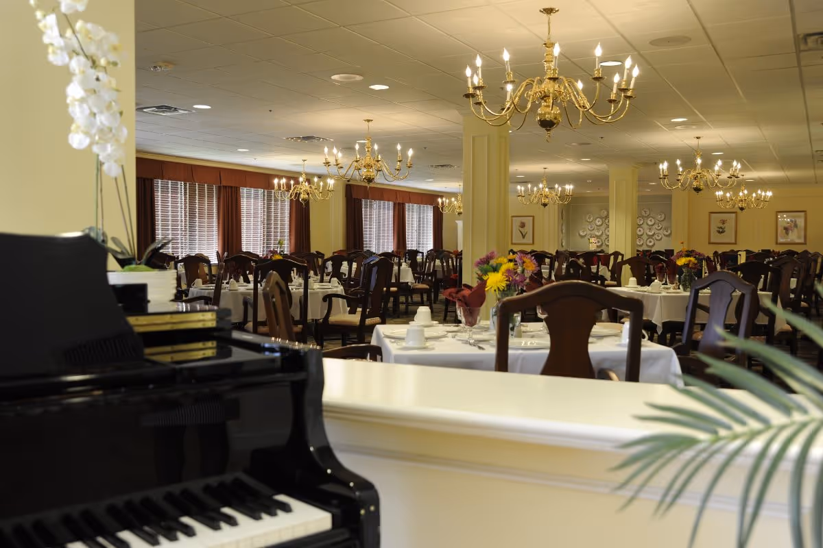 A spacious dining room with multiple tables covered in white tablecloths, set with cups, plates, and napkins. The room features elegant wooden chairs, chandeliers hanging from the ceiling, large windows with brown curtains, and a black piano in the foreground with a white orchid on top.