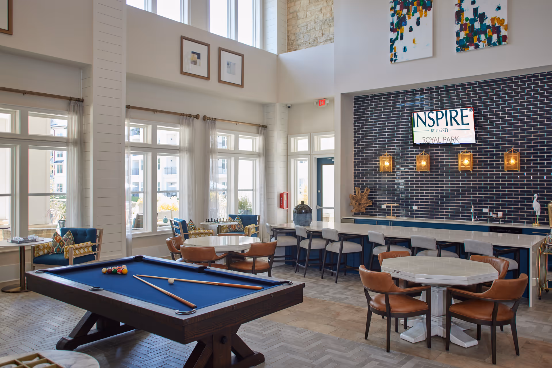 A spacious and well-lit common area in Inspire Royal Park featuring a blue pool table with cues and balls, several round tables with brown chairs, a long counter with bar stools, large windows with sheer curtains, and modern pendant lights hanging above the counter. The wall behind the counter is decorated with dark tiles and a sign that reads 'INSPIRE by Liberty ROYAL PARK'.