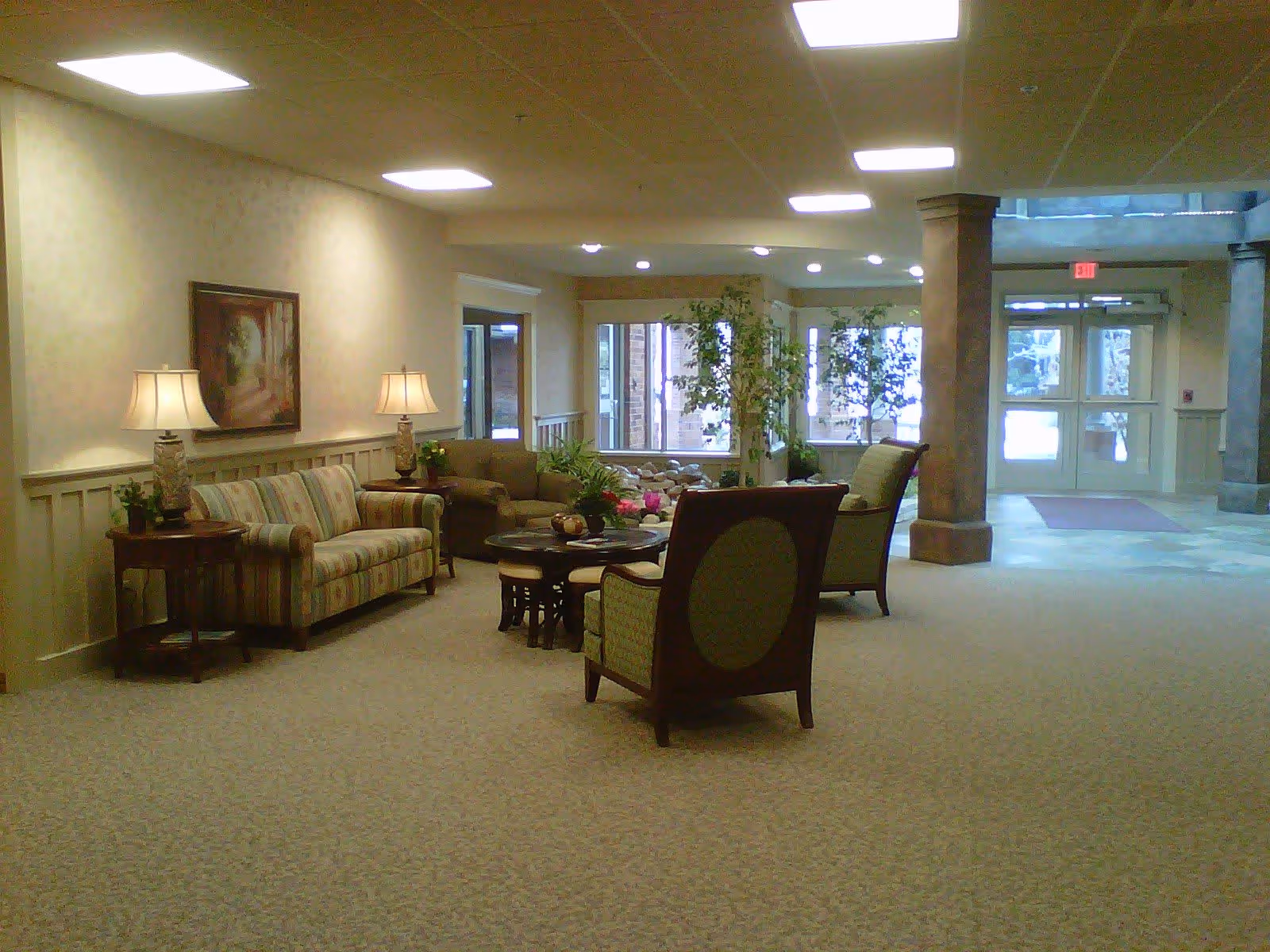 A spacious lobby seating area with sofas, armchairs, side tables and lamps, plants, and entrance doors in the background.