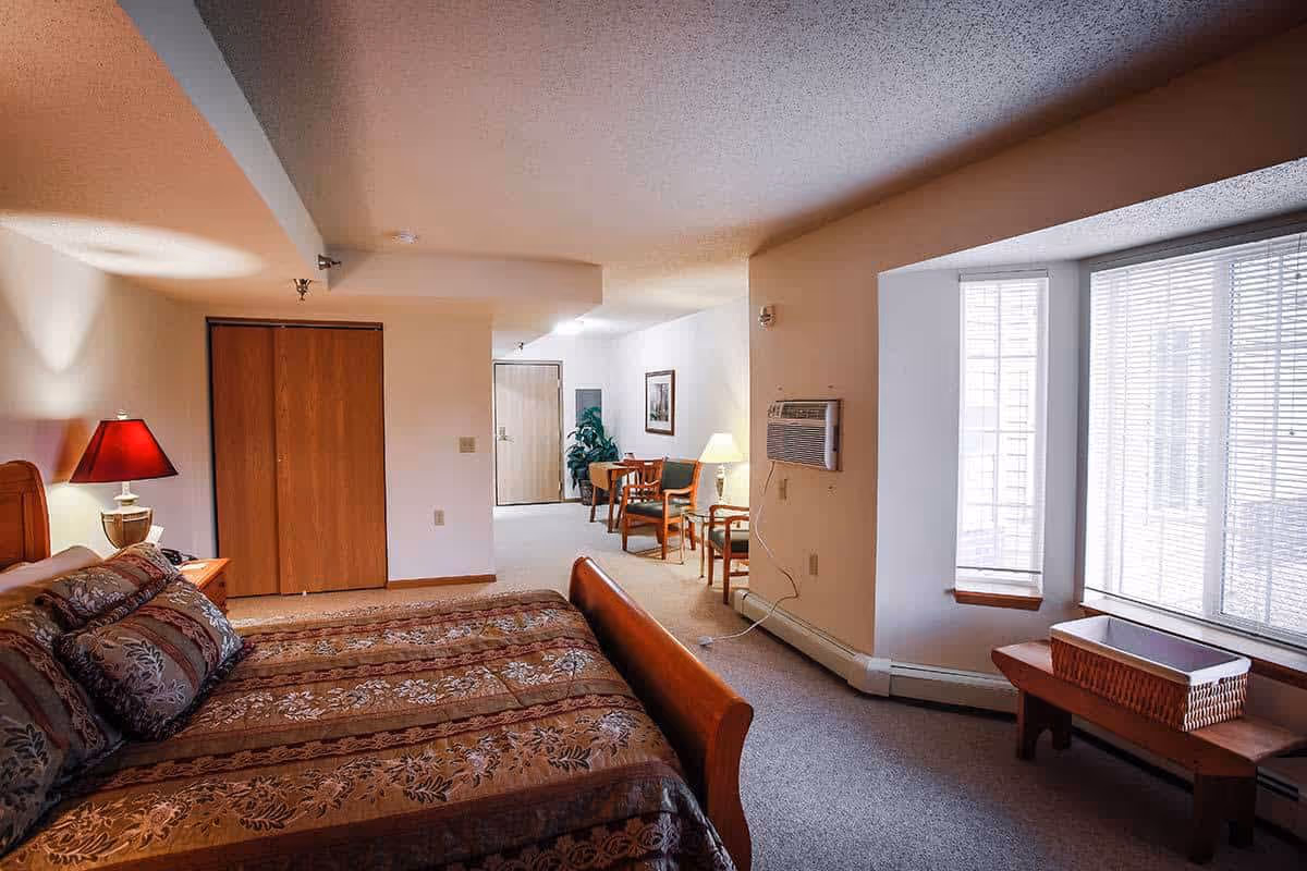 Large bedroom suite featuring a patterned bed, nightstand with a lamp, closet doors, a seating area with chairs and table, and a bay window with blinds.