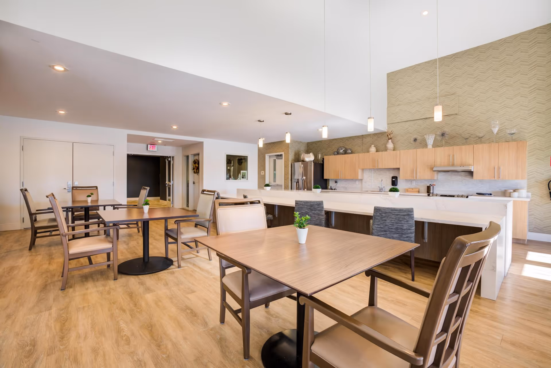 A bright and spacious dining area with wooden tables and chairs, small potted plants on each table, and a modern kitchen in the background featuring light wood cabinets, a white countertop, pendant lighting, and stainless steel appliances.