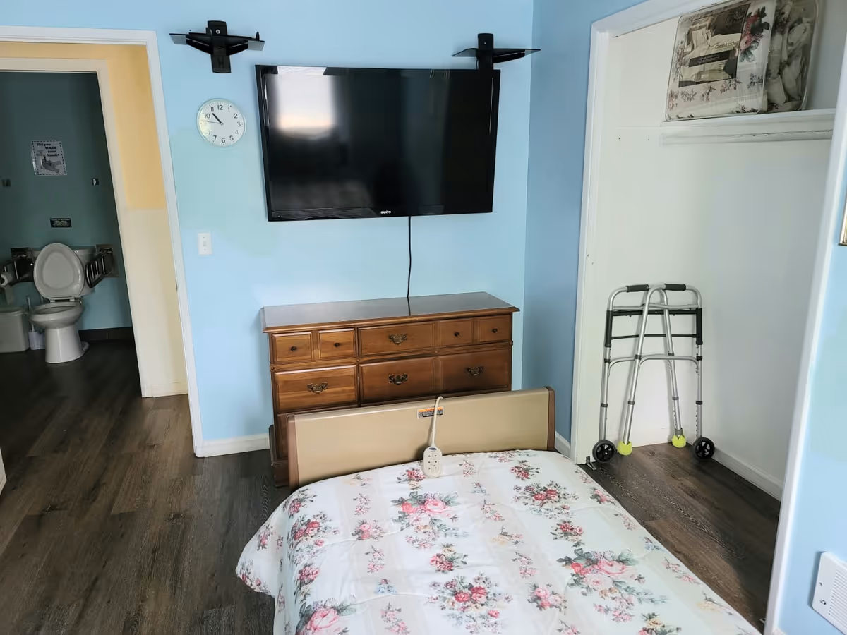 Small bedroom with floral bedding, a wooden dresser under a wall-mounted TV, a walker in an open closet, and a view into an adjacent bathroom.