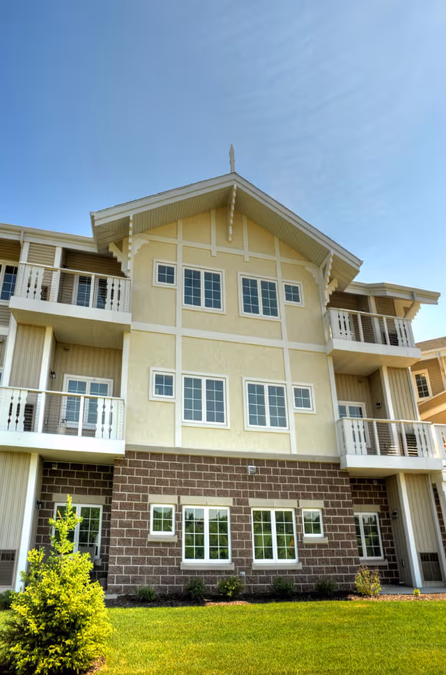 Front exterior of a three-story assisted living building with balconies, multiple windows, and a manicured lawn.