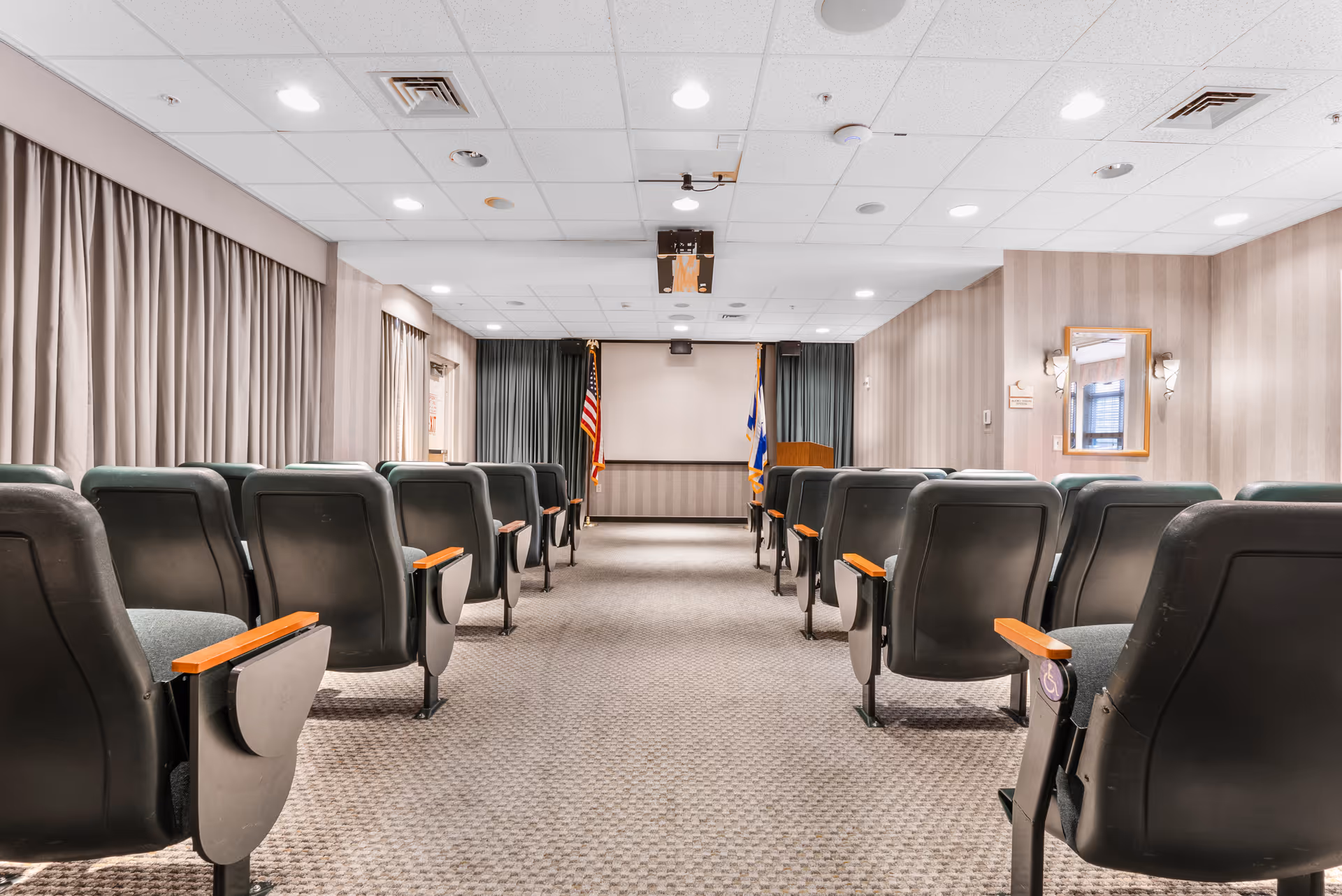 A small auditorium or meeting room with rows of green cushioned chairs with wooden armrests facing a podium and a projection screen. The room has beige patterned wallpaper, carpeted floor, and ceiling lights. Two flags are positioned on either side of the podium.