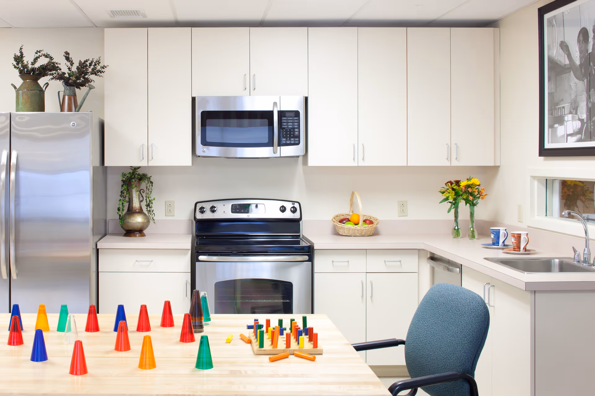 A bright kitchen with white cabinets, a stainless steel refrigerator, stove, and microwave. A wooden table in the foreground has colorful plastic cones and a pegboard game. There is a blue chair at the table, a basket of fruit on the counter, two mugs near the sink, and a framed black and white photo on the wall.